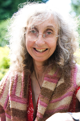 Head and shoulders photo of author smiling broady. She has a colourful shawl around her shoulders, pink and brown. Her hair is shoulder length and irradiated by sunlight.