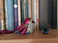 Colour photo of bookshelf with two little birds, one a Xmas decoration and one glass. The birds are looking at each other. Behind them a row of book spines, various colours.