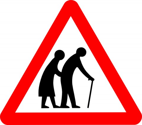 Elderly people crossing road sign, depicting two old people. The old man is in front with a stick. The old woman stoops alongjust behind him. It's quite a sexist sign!
