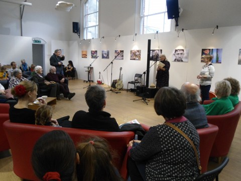 Annie Fisher reading with microphone to an audience in a large hall, comfy seats in a big semi circle and light from big overhead windows. Behind her, along the white wall a striking exhibition of photographs.