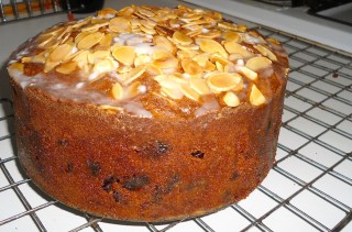 Picture shows a large cake on a baking tray. It's some kind of A Deep dark golden sponge, clearly containing fruit and on the top flaked almonds and drizzled white icing.