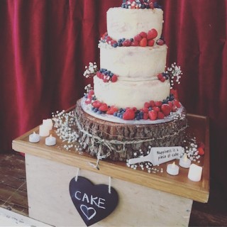 Full colour photo of wedding cake surrounded by soft fruit, strawberries, blueberries, raspberries. The cake slice has lettering on it that reads Happiness is a piece of cake.