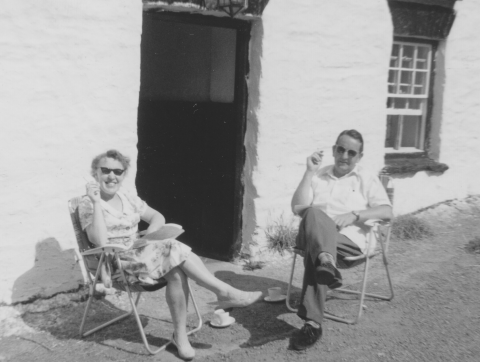 Monchrome photo of a youngish couple sitting on two folding chairs outside the door of a holiday cottage. Each has a cup and saucer on the ground beside the chair. Each has a cigarette in the right hand. Both are wearing dark glasses and smiles. It must be a very warm day because the short haired guy (my father) is wearing sunglasses, and so is my mother. This picture was taken in South Wales.