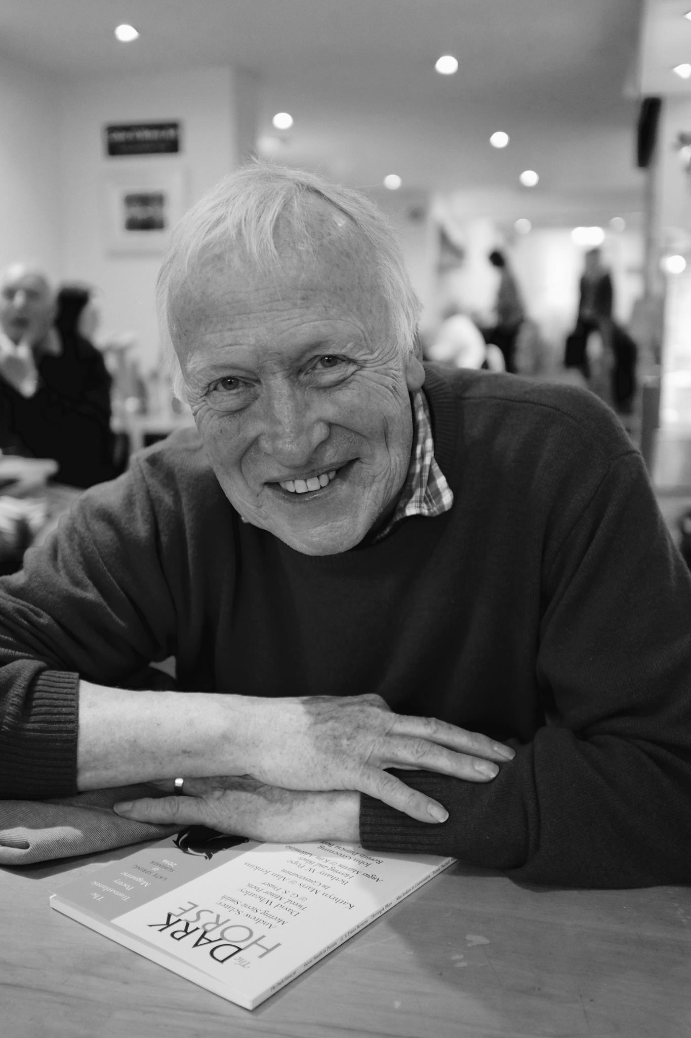 Photo of Andrew leaning forward over a table, his arms folded on the surface. A big smile, white teeth, no glasses. He is warring a casual pullover and a collared t-shirt.
