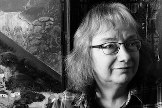 Black and white photo of author looking slightly to the right with a relaxed friendly expression. She has hair that just touches her shoulders and wears glasses and an open necked shirt. Behind her could be a window, could be a picture...