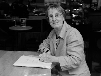 The author is pictured in monochrome sitting at a cafe table, pen in hand and a manuscript in front of her. She is looking at the photographer with a mischievous smile. She has short hair and glasses and is wearing a smartish jacket.
