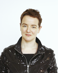 A full colour head and shoulders picture with the face square on looking the viewer right in the eye. She is smiling very slightly. A youngish woman with a boyish haircut, a clear open forehead, dark brown hair and eyebrows, blue eyes. She is wearing a glittery jacket, zipped open at the neck, and below it a plain black t-shirt with a straight neckline. No  jewellery of any sort.